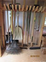 Wood and metal handle garden tools including shovels, rakes, broom, pitch fork hung on wooden rack inside shed or garage.