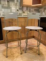 Two grey kitchen stools with metal frames, round cushioned seats, rectangular backrests with handle cutouts in a kitchen corner.