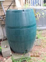 Single green plastic rain barrel outdoors with attached white downspout and spout near base