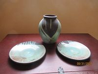 Photo showing the Raku vessel centered between the two matching platters on a wooden surface under warm lighting, highlighting glaze colors and textures.