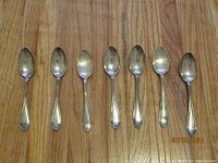 Seven sterling silver teaspoons lined up evenly, showing their front sides with smooth, simple handles and rounded bowls.