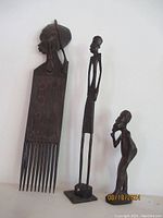 Photo of three vintage African hand carved wood items: a large wall hanging comb with woman figurine, a tall standing man statue, and a smaller tribal art figurine.