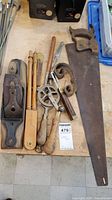 Photo showing an assortment of antique woodworking tools including saw, hand planer, brace drill, and dividers on a wooden surface.