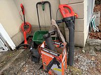 Overview of all items including leaf blower in box, weed wacker, hedge trimmer, and spreader, shown outdoors on gravel and concrete surface.