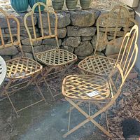Four vintage wrought iron folding patio chairs set outdoors on stone and leaf-covered ground, showing weathered paint and rust spots.