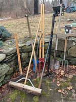 Full view of all yard tools including scythe, earth packer, pick axe, broom, and various hand tools standing outdoors by stone wall