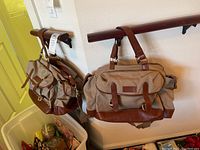 Two Brooks Brothers tan canvas and brown leather shoulder bags hanging on a wooden rail, showing the overall style and size of the bags.