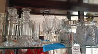 Wide view showing multiple crystal candle holders, stemware, a pitcher, and a large jar with lid on glass shelving.