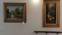 Two framed oil paintings hanging on wall, one of a countryside bridge scene, the other a still life with fruit and bottle.