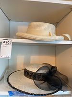 Two ivory woven ladies hats on shelf: one with black lace band and large black mesh bow, the other with beige band and ribbon.