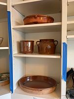 Four pottery pieces displayed on shelves including a large round pottery platter at the bottom, two medium-sized pots with handles on the middle shelf, and a shallow bowl-shaped platter at the top.