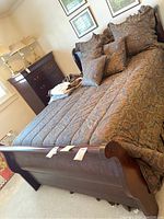 Full view of queen size sleigh bed with bedding and pillows in traditional paisley pattern, placed in bedroom with chest of drawers and lamps in background.
