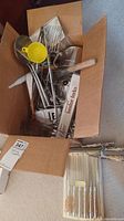 Open cardboard box filled with assorted kitchen utensils including metal spoons, ladle, whisk, yellow measuring scoop, strainer, and boxed fondue forks set.