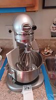 Front and partial top view of silver KitchenAid stand mixer with whisk attachment and stainless steel bowl attached.