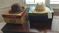 Two hats displayed on a wooden table near a window: Brown Stetson fedora on its box and beige Churchill straw hat on its box.