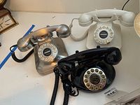Three vintage rotary dial telephones: black, silver, and cream-colored models with coiled handsets shown on a white surface.