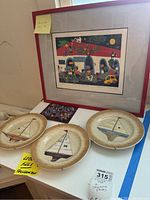 Photo showing three collector plates each with sailboat designs and the framed colorful signed wall art behind them.