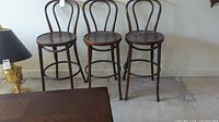 Three wooden bar stools standing side by side against a wall on carpeted floor. All have bentwood curved backs, round seats, four legs, and footrests.