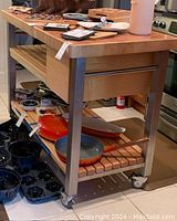 Front/right three-quarter view of butcher block island showing maple top, stainless legs, drawer front panel and slatted shelf with cookware.