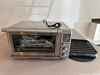 Front and side view of stainless steel Breville convection smart toaster oven with control panel showing knobs and digital display. Accessories are placed next to oven, including black drip pan and grill rack wrapped in plastic.