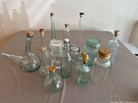 Overview of 13 glass bottles with various shapes and cork or clip-top stoppers on a gray tablecloth.