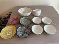 Image showing 11 ceramic bowls on table: 5 multicolored patterned shallow bowls, 4 plain white bowls, and 2 larger white basket-weave style bowls.