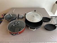 Full view of the cookware group on a table with multiple frying pans and lids, including a red pan and cast iron skillets.