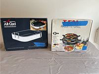 Two boxed items: All-Clad rectangular roasting pan with rack and T-fal Raclette Grill, both in original packaging with stickers indicating the lot.