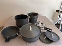 Photo of a collection of black pots and pans with lids laid out on a table, showing different sizes and handle types.