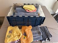 Plastic bin filled with folded and stacked dish towels in various colors and patterns, some with floral embroidery designs such as lavender and other flowers, others plain or striped.