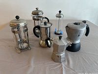 Photo of six coffee brewing devices including four French presses and two stovetop coffee pots arranged on a white surface against a plain background.