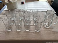 Set of 12 smaller clear drinking glasses and one large beer stein on a beige cloth.