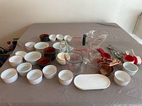 Wide view of multiple kitchen items including Pyrex measuring cups, Apilco porcelain ramekins and bowls, copper measuring cups, clay bowls and measuring spoons spread on table.