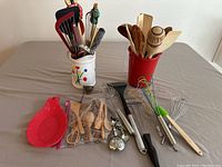 Photo showing a variety of kitchen utensils: wooden spoons and spatulas in a red ceramic holder, metal whisks with wood and silicone handles, plastic spatulas, various metal measuring spoons, and a garlic press, with some small wooden utensils in a clear plastic bag.