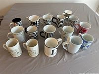 Group of 15 ceramic coffee mugs arranged on a table showcasing various designs including black and white patterns and colored mugs.