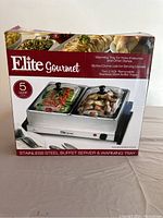 Box front shows the Elite Gourmet branding and highlights features: warming tray for hors d'oeuvres and other dishes, two 2.5 quart removable stainless steel buffet trays, slotted dome lids for serving utensils.
