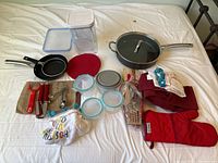 Overview of all cookware and kitchen gadgets laid out on a bed, showing frying pans, containers, towels, mitt, utensils, and more.