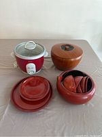 Red Oster rice cooker with white handles, control buttons, and glass lid, placed beside three red plates, a large red bowl with nested small bowls inside, and a wooden bowl with lid.