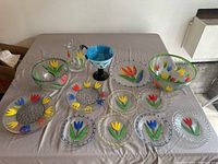 Overall set of colorful hand painted Kosta Boda glass dinnerware on display cloth including bowls, plates, pitcher, and decorative stemmed bowl.