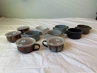 All stoneware items arranged on white fabric: one larger bowl, six brown soup bowls with handles and lids, and four grey/tan soup bowls without lids.