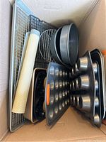 Overhead photo of various oven and baking items placed inside a cardboard box, including metal cooling racks, multiple muffin trays of varying sizes and cavity counts, several round baking pans, a large rectangular baking pan, and a white silicone baking mat.