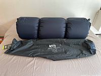 Inflatable sleeping pad partially inflated, showing dark fabric and included storage bag with REI Camp Bed 3.5 logo.