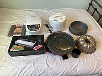 Wide view of all the items in the lot arranged on a bed including white Tiger rice cooker, vacuum sealer box, floral white electric cooker, and bakeware pans.
