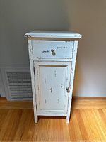 Front view of small white rustic bathroom cabinet showing drawer and cupboard door with distressed paint and scuff marks.