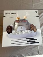 Box showing the product in use with marshmallows being roasted over the central heating element, surrounded by marshmallows, chocolate, and crackers.