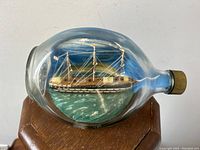 Front view showing the schooner ship model inside the clear glass bottle with painted ocean and sky background.