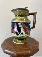 Front view showing boy and dog relief on the pitcher jug with mottled green and blue glaze and metal lid.