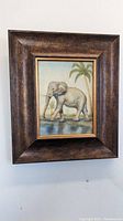 Framed art print featuring an elephant walking by water with palm trees in the background, showing overall condition and frame details.