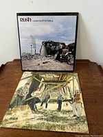 Two vinyl LP records, 'A Farewell to Kings' by Rush displayed upright, and 'The Best of The Guess Who' with band photo cover laying flat on wooden surface.