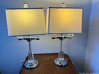 Pair of solid crystal glass table lamps illuminated on a wooden table with beige rectangular fabric shades. Lamps have chrome bases and clear crystal tapered stems.